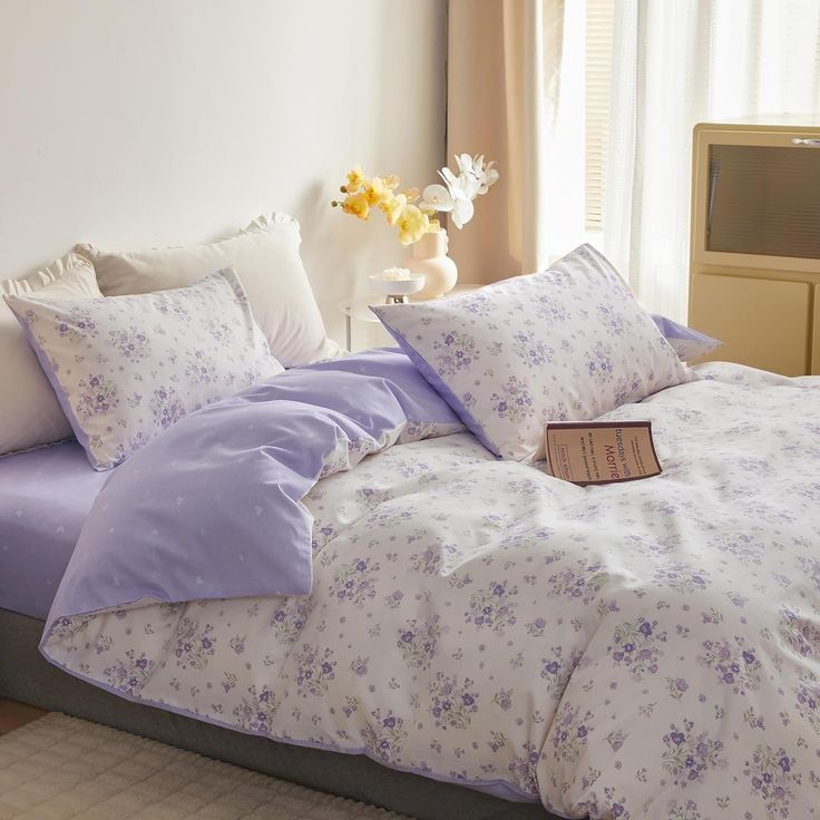 a bed with purple and white comforters in a bedroom next to a vase filled with flowers