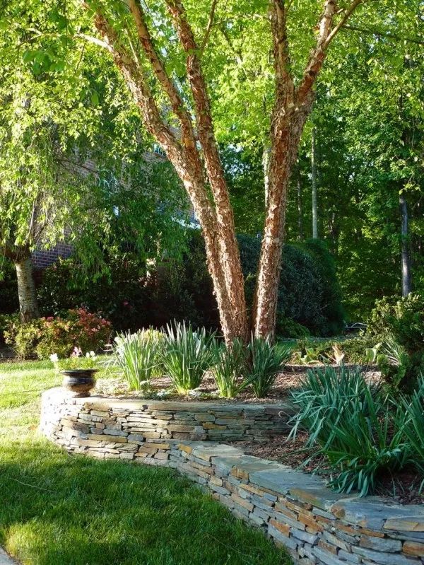 a stone wall in the middle of a grassy area next to a tree and shrubbery