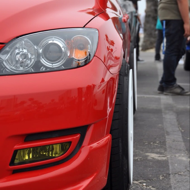 the front end of a red car parked in a parking lot with people walking by