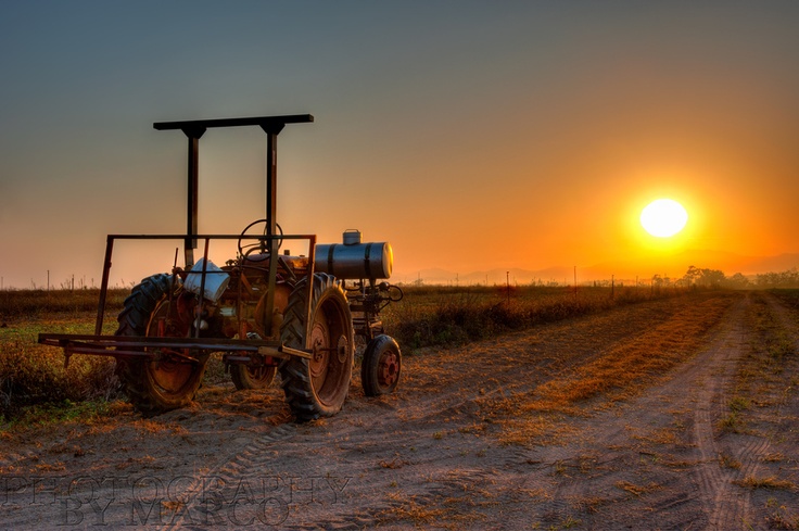 an old tractor sitting in the middle of a dirt road with the sun setting behind it