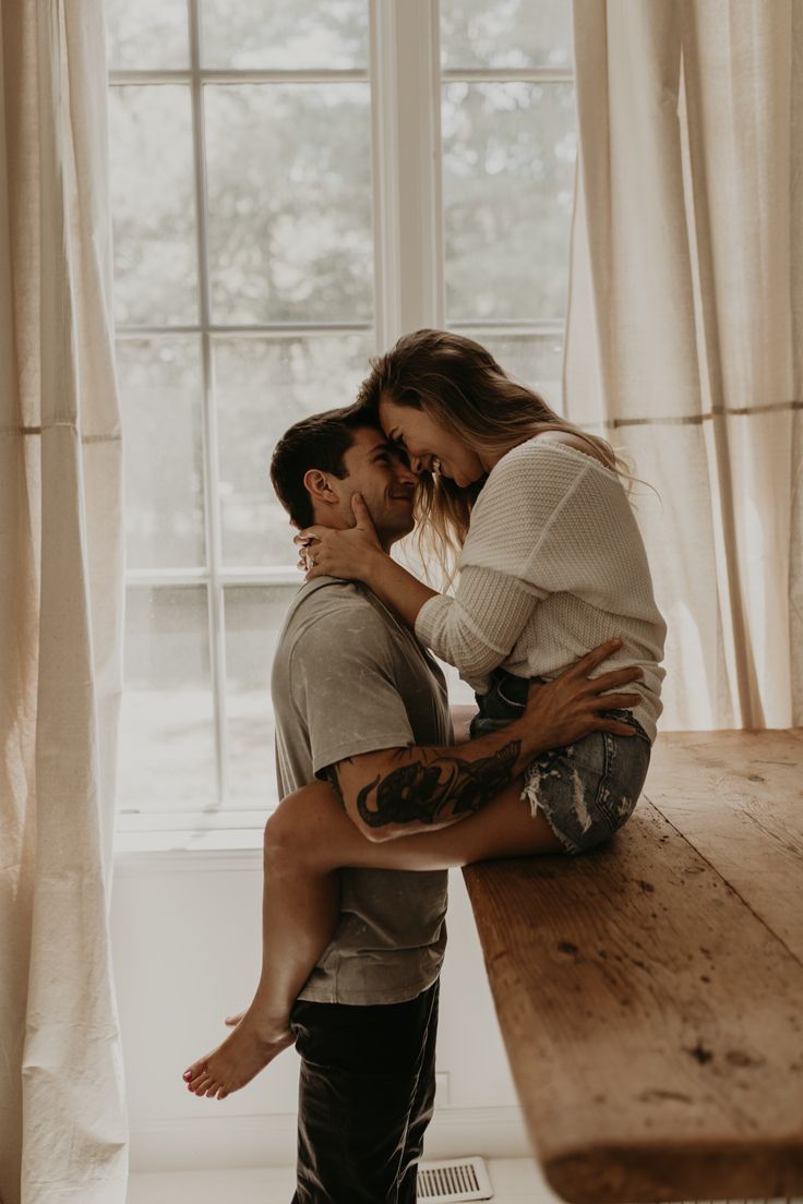 a man holding a woman in his arms while standing next to a table near a window