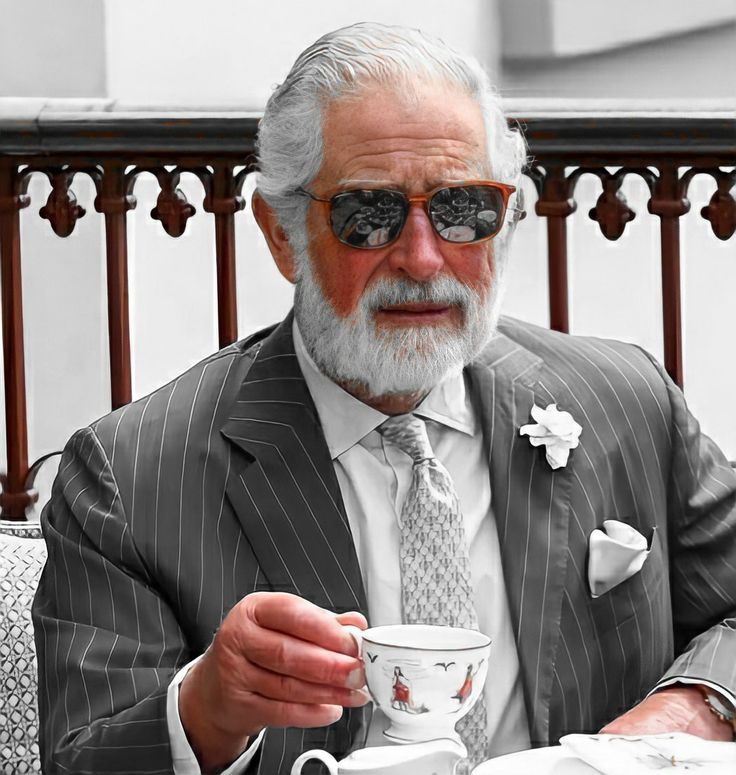 an older man in a suit and tie holding a coffee cup while sitting at a table