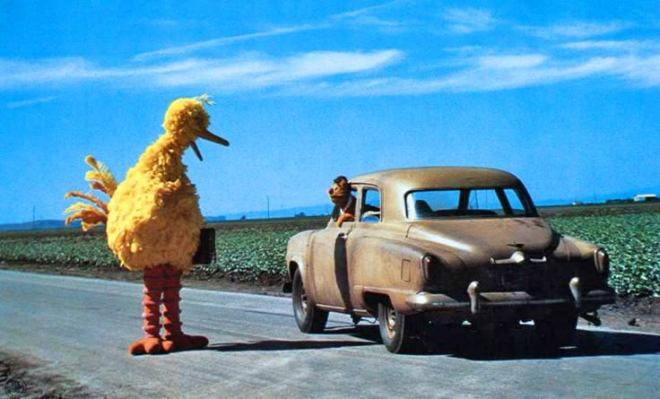 an old car driving down the road with a large bird standing next to it