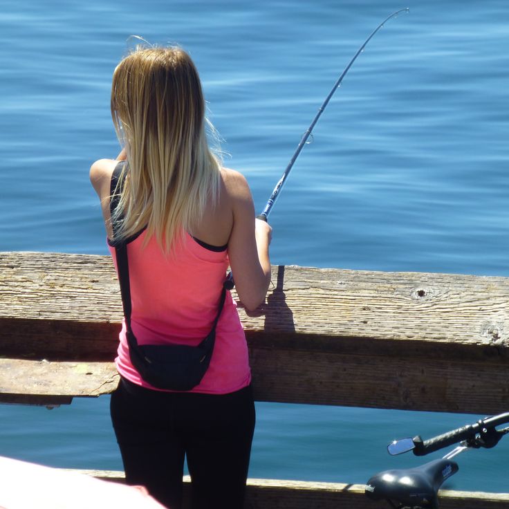 the woman is fishing on the pier by the water while wearing a pink tank top