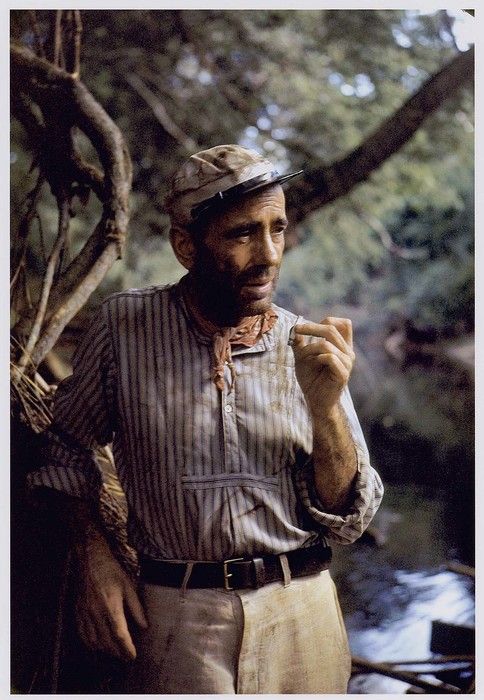 a man standing next to a tree holding something in his hand while wearing a hat