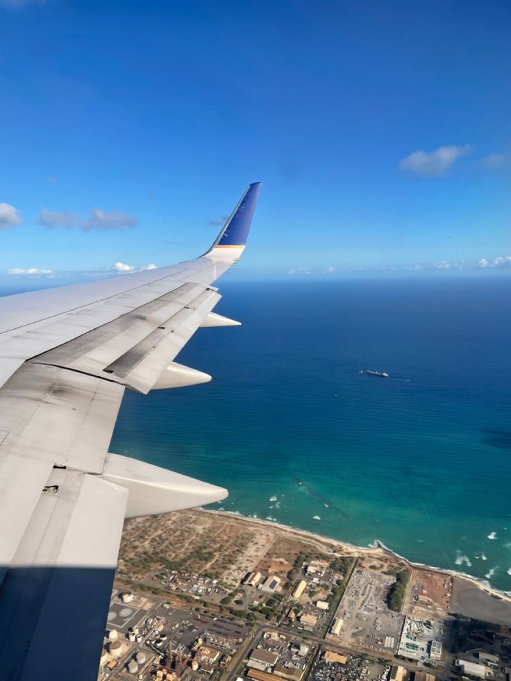 the wing of an airplane flying over a city next to the ocean on a sunny day