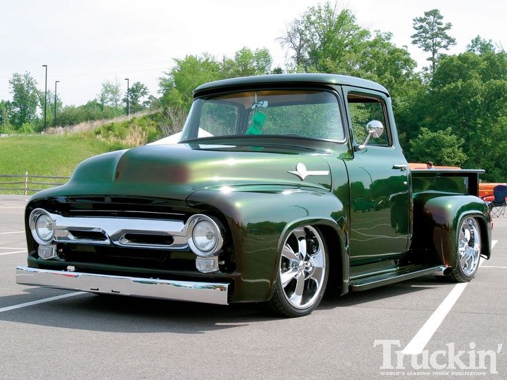 an old green pickup truck parked in a parking lot with trees and grass behind it