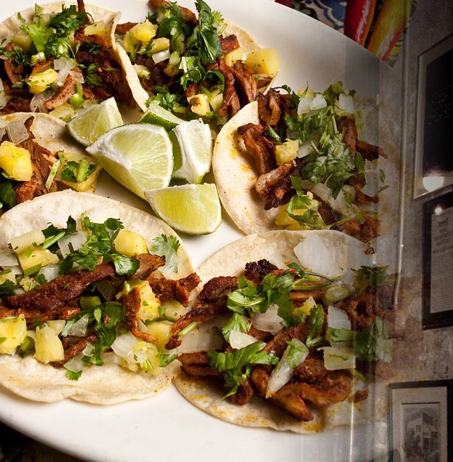 several tacos on a white plate with limes and other ingredients in the background