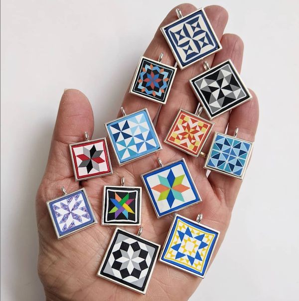 a hand is holding nine small square earrings in different colors and designs, each with an individual's finger