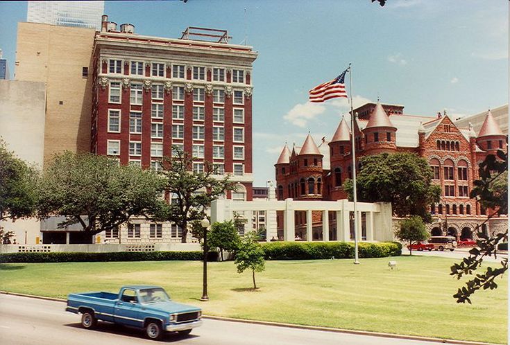 an old blue car is parked in front of a large building and flagpoles