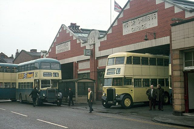 two double decker buses parked in front of a building with people standing around it and one bus is yellow