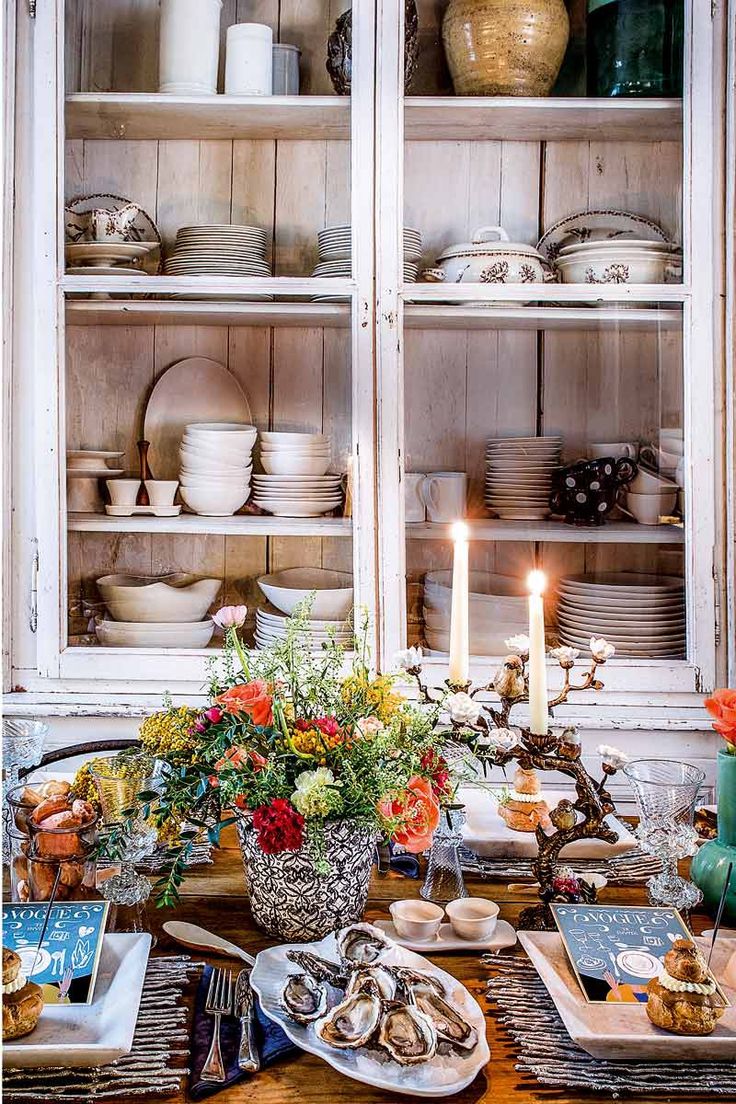 a dining room table with plates and flowers on it, candles in the center surrounded by china cabinets