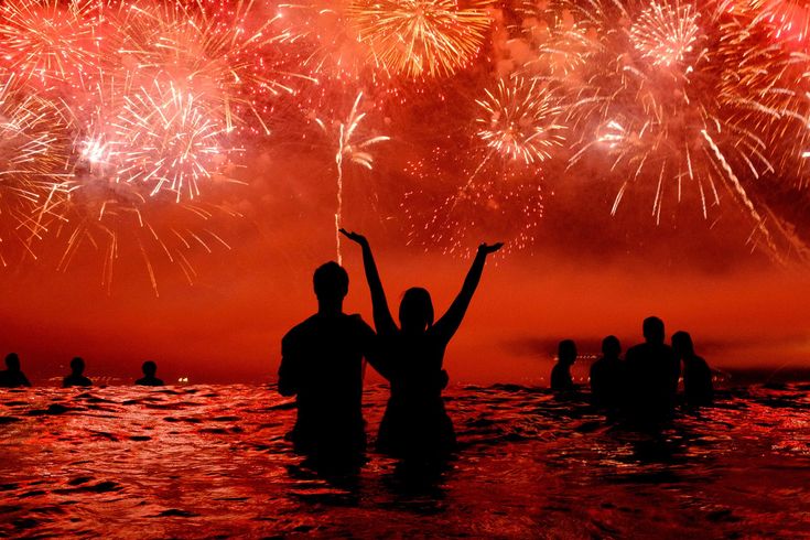 two people are standing in the water with their arms up as fireworks go off behind them