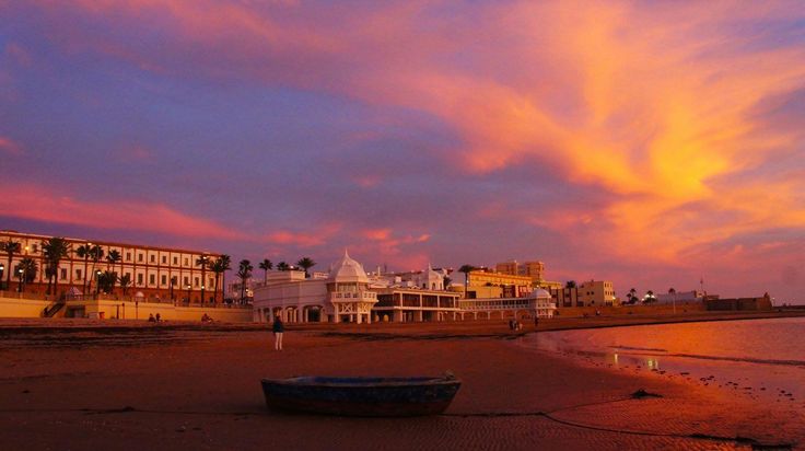 a boat sitting on top of a sandy beach under a colorful sky at sunset with buildings in the background