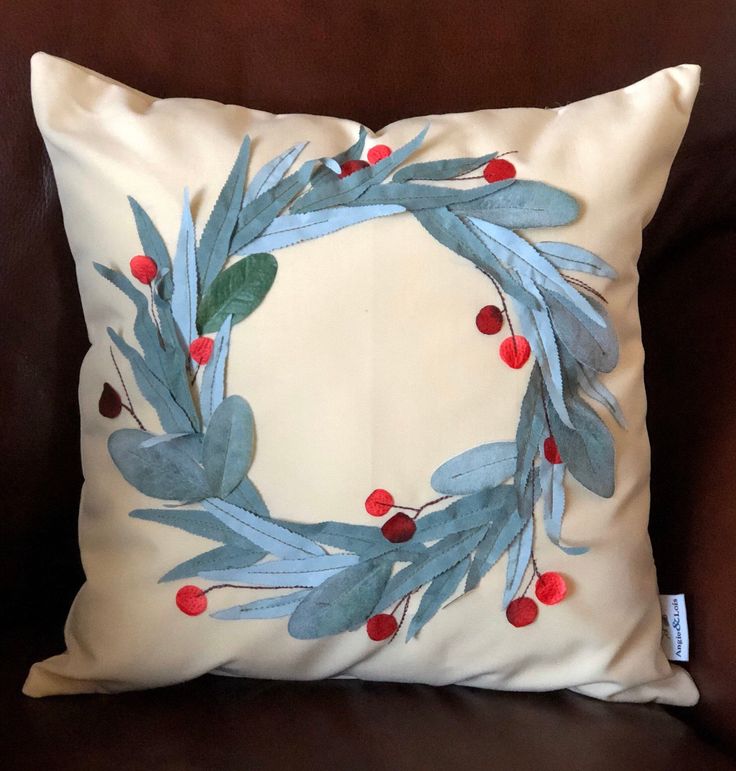 a white pillow with blue leaves and red berries on it, sitting on a brown couch