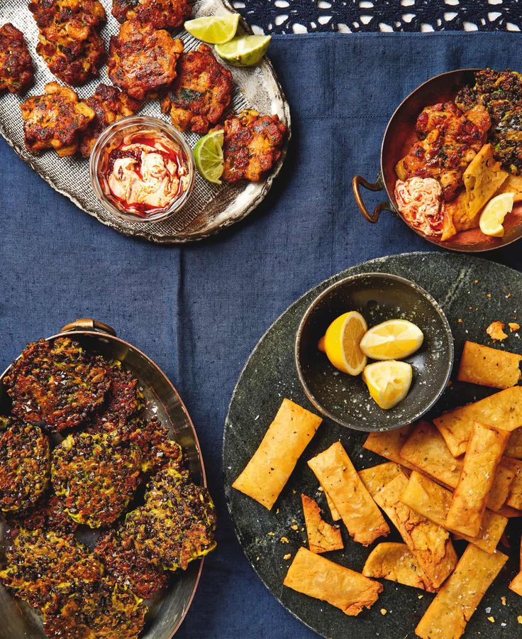 several plates of food on a blue table cloth with lemon wedges and pita chips