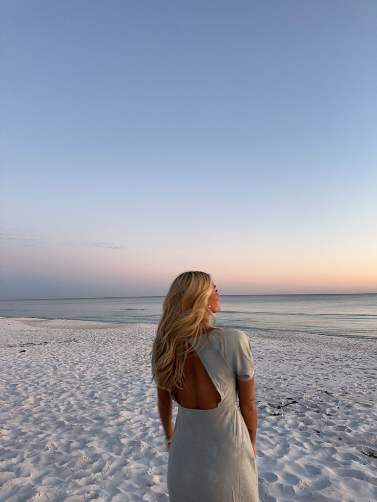 a woman standing on top of a sandy beach next to the ocean with her back turned