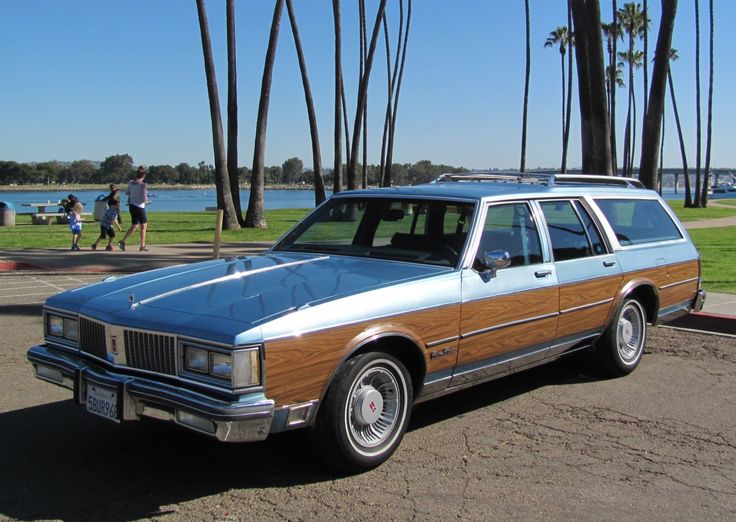 an old station wagon is parked in front of some palm trees and people walking by