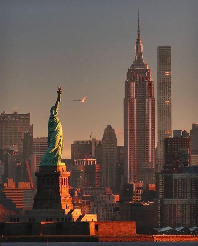 the statue of liberty in new york city is lit up at sunset with skyscrapers behind it