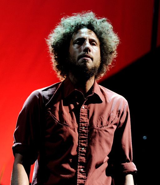 a man with curly hair standing in front of a red background wearing a brown shirt