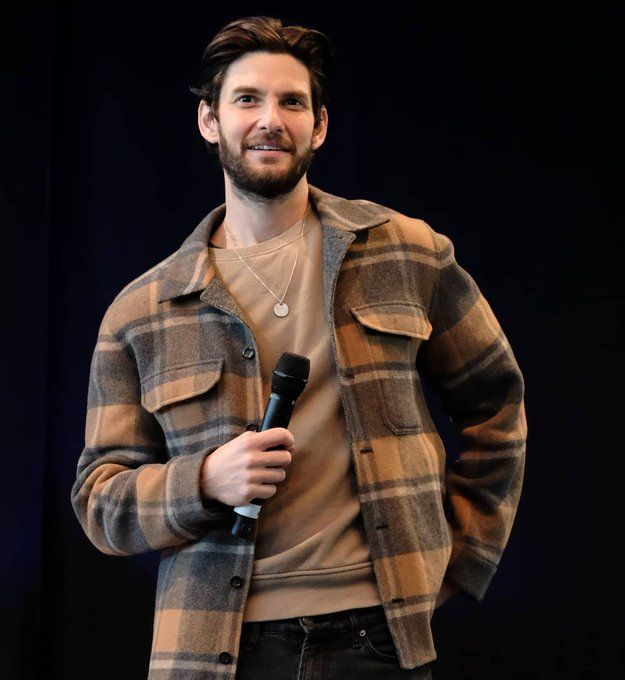 a man standing in front of a black background holding a microphone and wearing a plaid jacket