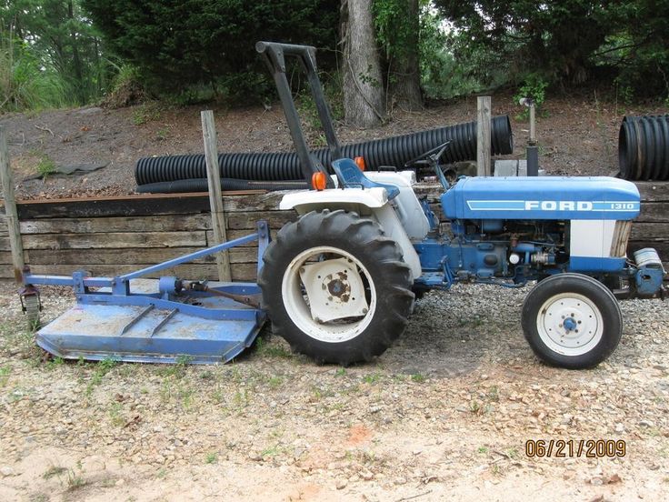 an old blue and white tractor sitting on the ground next to a wooden fence with trees in the background