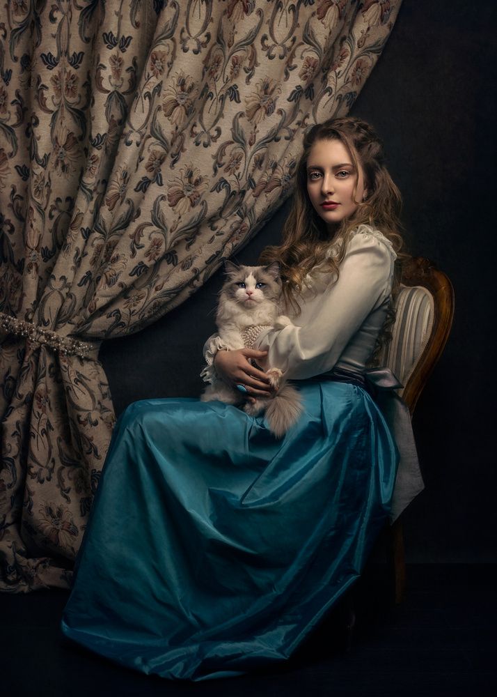 a woman sitting on a chair holding a cat in front of a window with curtains