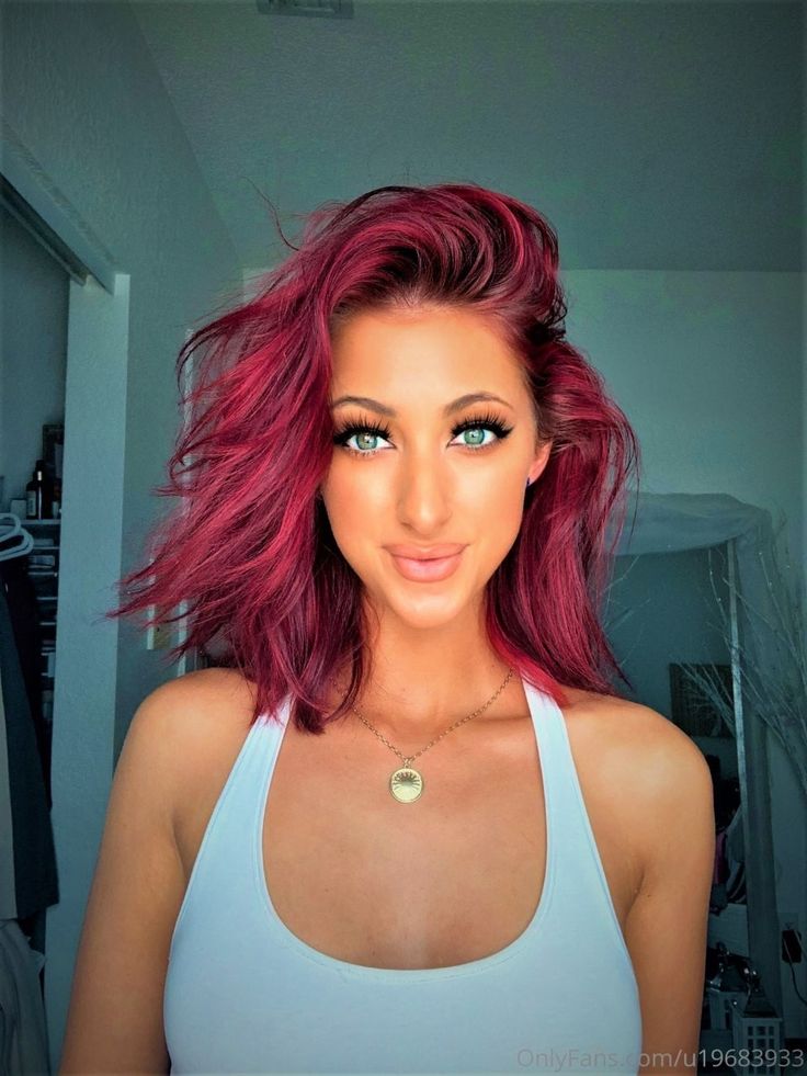 a woman with pink hair and green eyes wearing a white tank top is smiling at the camera