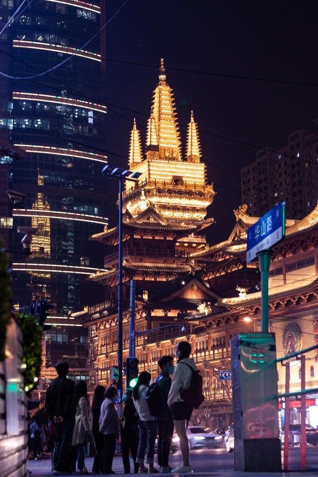 many people are standing on the sidewalk in front of some tall buildings at night time