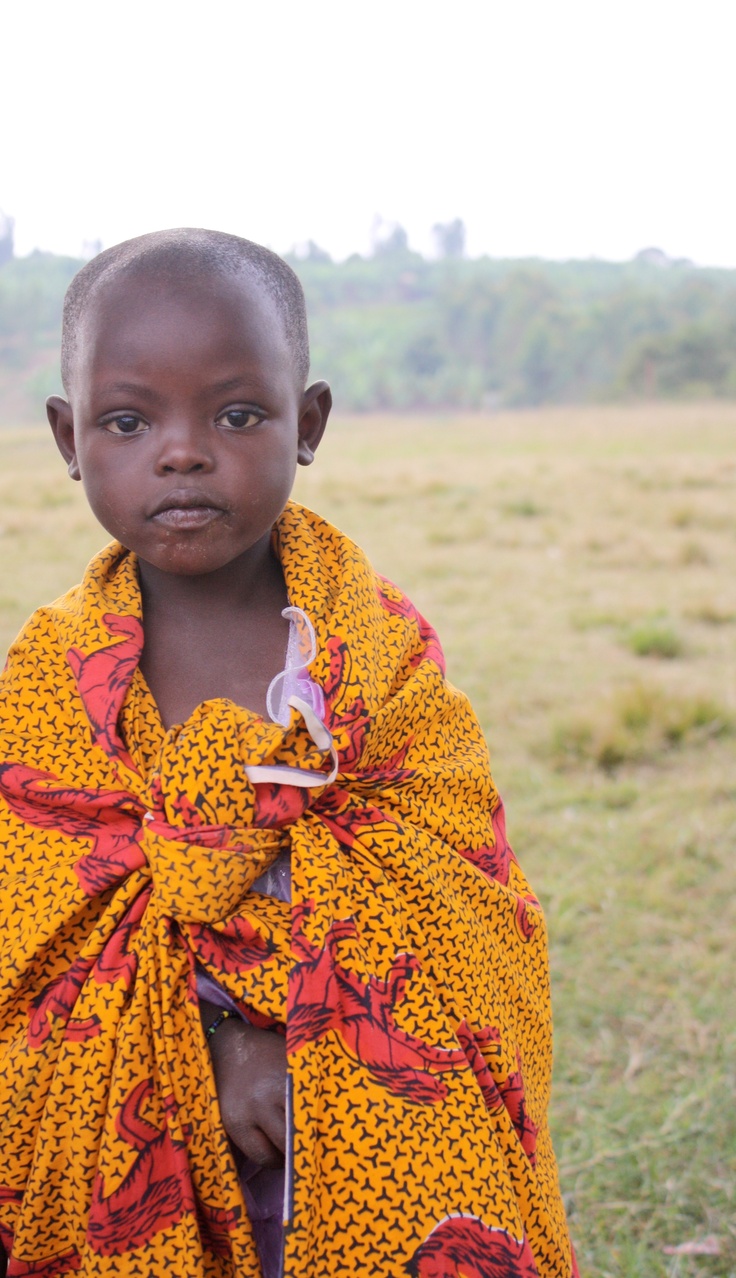 a young boy wearing a yellow and red shawl standing in a grassy field with trees in the background