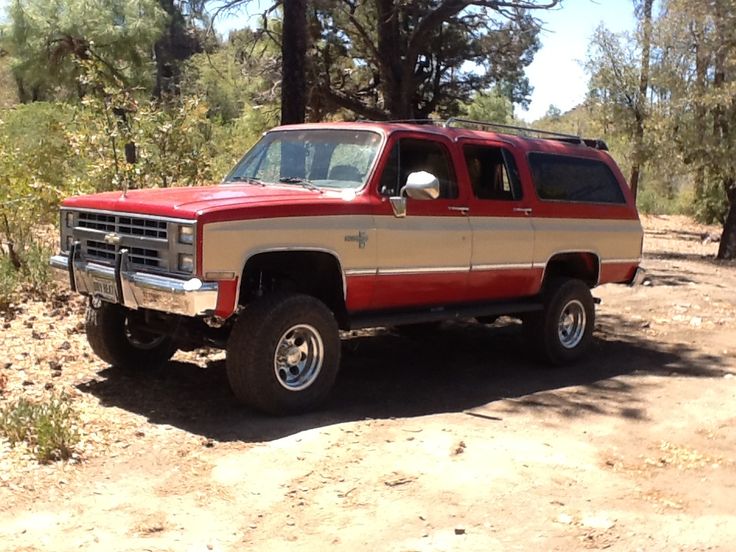 a red and tan truck is parked in the dirt near some trees on a sunny day