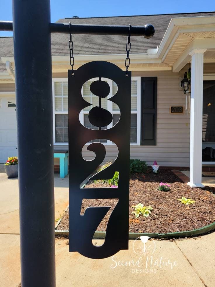 a metal house number sign hanging from a black pole in front of a white house