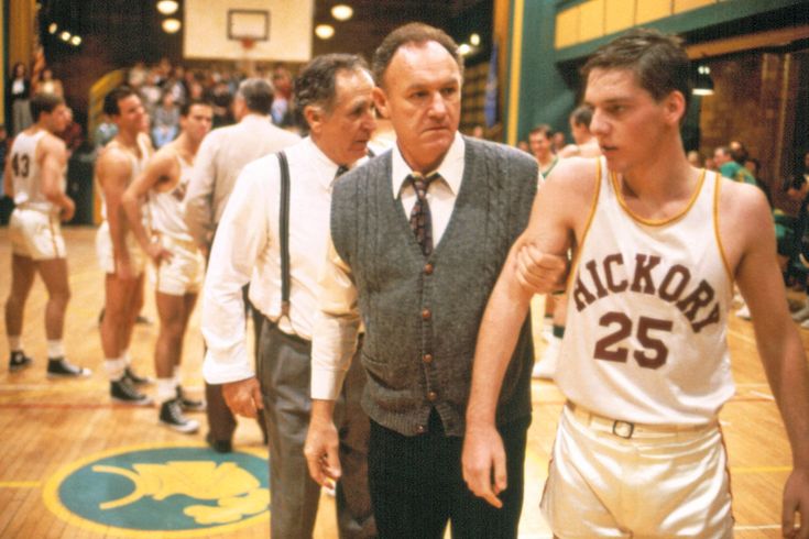 two men walking on a basketball court with other men in the background wearing sports uniforms