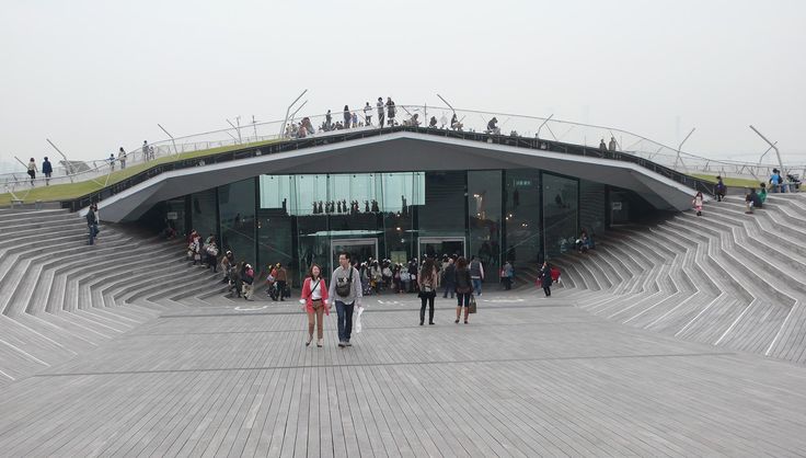 people are walking around in front of a building with an open roof and grass on top