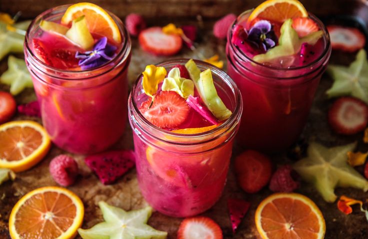 three jars filled with fruit and garnish on top of a wooden table next to sliced oranges