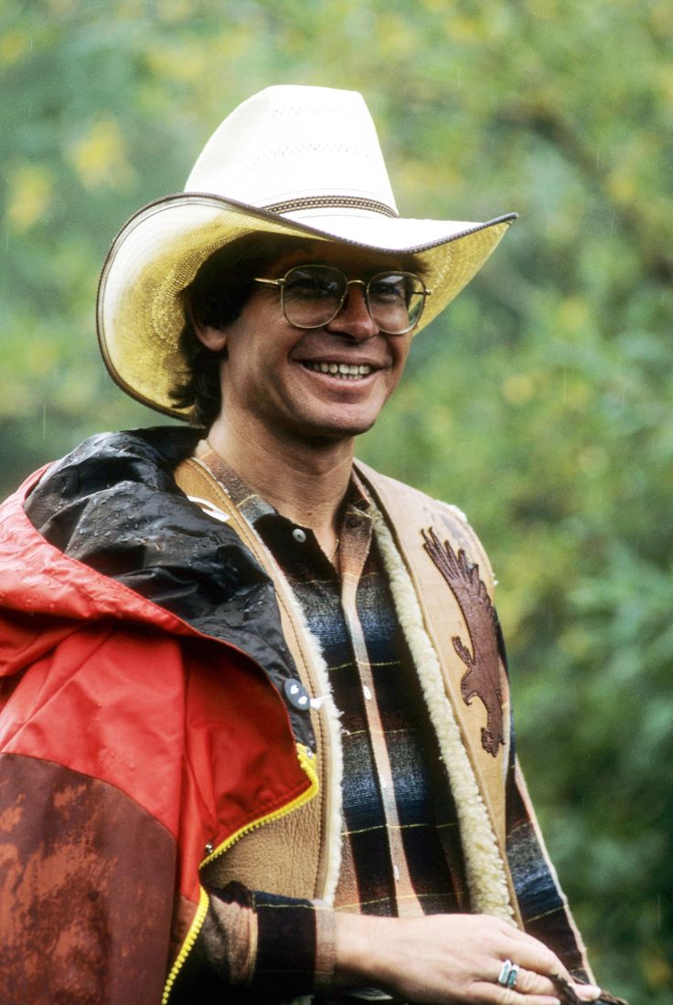 a man wearing a cowboy hat and glasses smiles at the camera while standing in front of trees