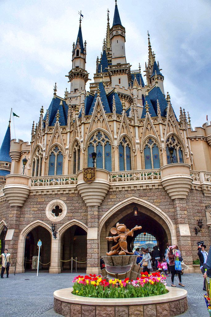 the entrance to sleeping beauty castle with flowers in front and people walking around it on a cloudy day