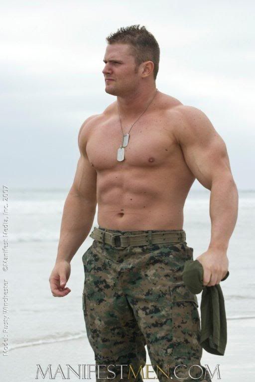 a shirtless man standing on the beach with his hands in his pockets and looking off into the distance