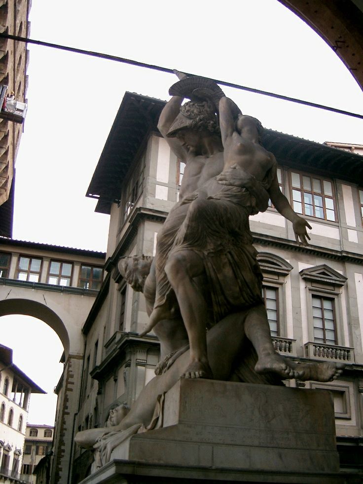 a statue in front of a building with an arch above it and buildings behind it