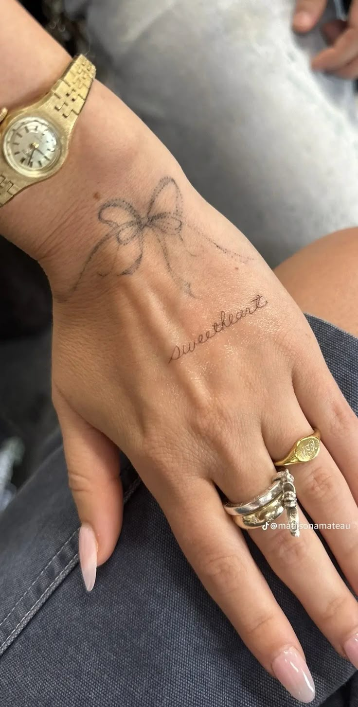 a woman's hand with a bow tattoo on her left wrist and gold ring