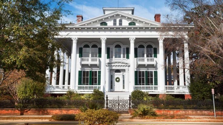 a large white house with green shutters on the front and second story, surrounded by trees