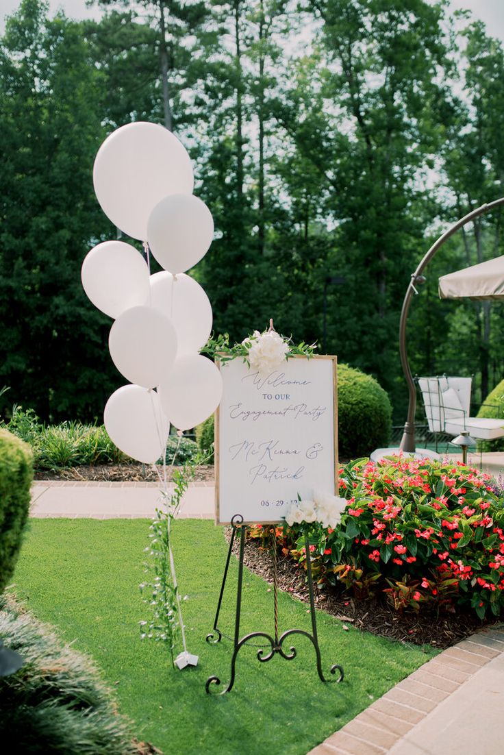 a sign with balloons attached to it sitting in the grass next to flowers and trees