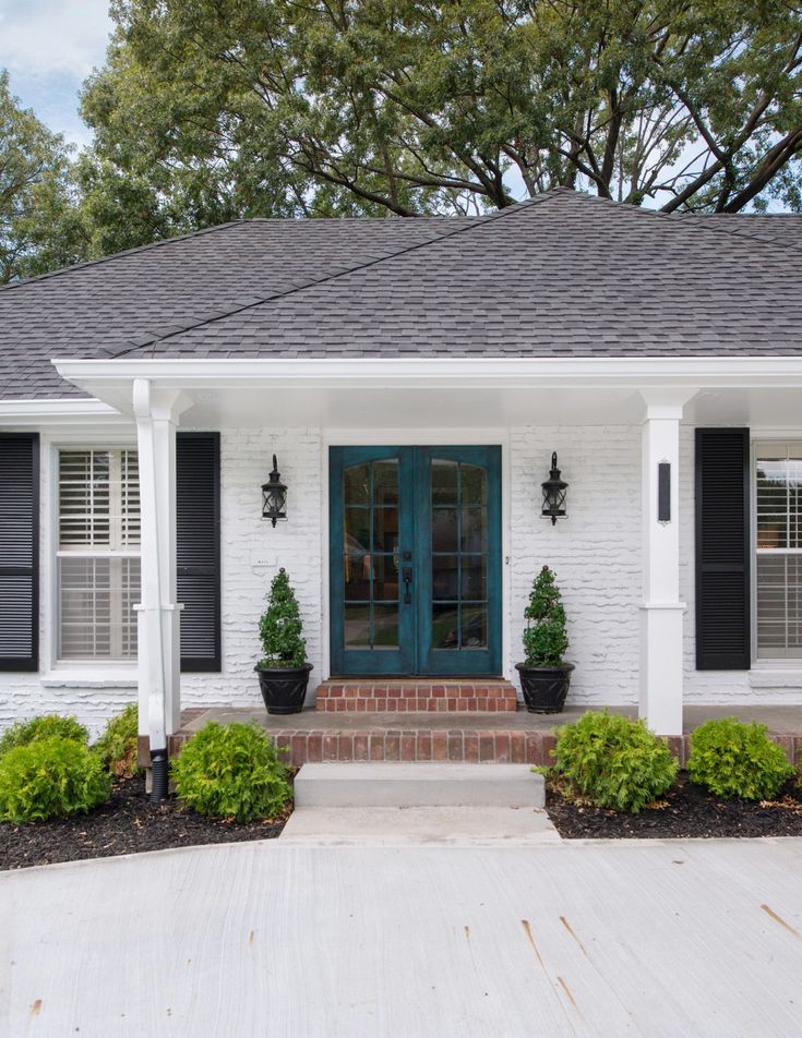 a white brick house with black shutters and green plants on the front door is shown