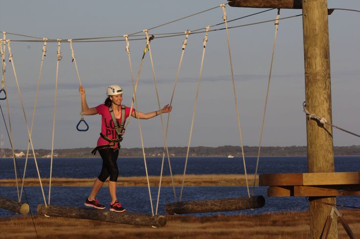 a woman walking across a wooden bridge with ropes above her and water in the background