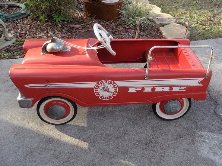 an old fashioned fire engine pedal car is parked on the sidewalk in front of some bushes