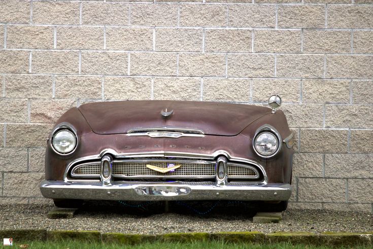 an old car parked in front of a brick wall with grass growing on the ground