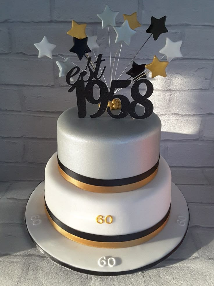 a white cake with black and gold decorations on it's tiers is displayed in front of a brick wall