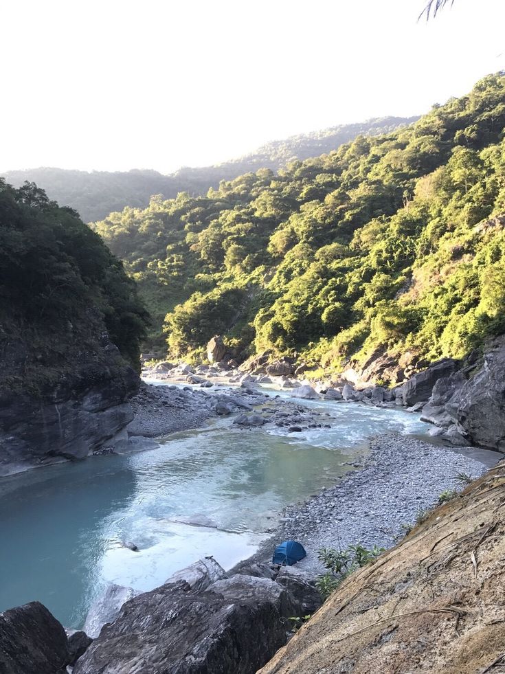 a river flowing through a lush green forest covered hillside next to a rocky cliff side