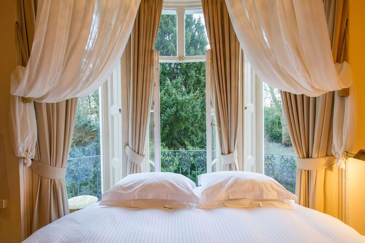 a bed with two pillows on top of it in front of a window that has curtains and drapes