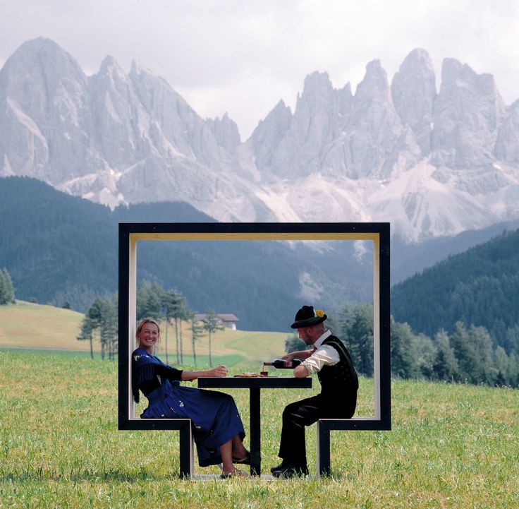 two people sitting at a table in the middle of a field with mountains in the background